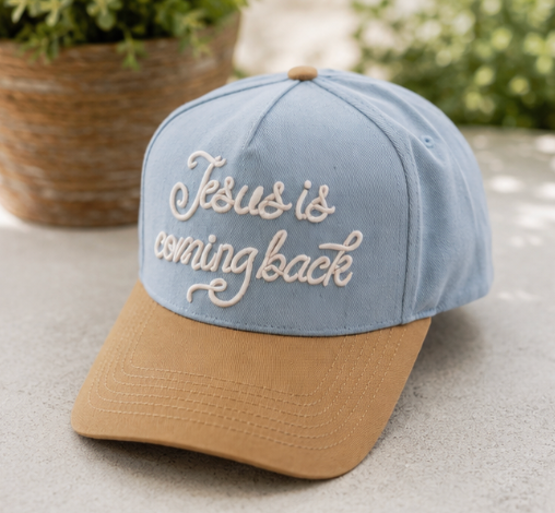 A two-tone baseball cap with a light blue crown and a tan visor, featuring the phrase 'Jesus is coming back' embroidered in white script on the front. The cap is photographed outdoors on a light gray stone surface with blurred green plants in the background.