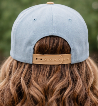 Back view of a person wearing a light blue baseball cap with a tan adjustable strap, shown outdoors with wavy brown hair.