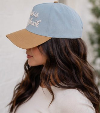 A woman in profile wearing a light blue baseball cap with a tan visor and embroidered text on the front, photographed against a soft light background.