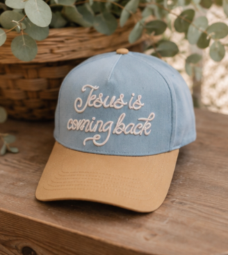 A two-tone baseball cap with a dusty blue crown and a tan visor, featuring the phrase 'Jesus is coming back' embroidered in white script across the front. The cap is styled on a rustic wooden surface with a wicker basket and eucalyptus leaves in the background.