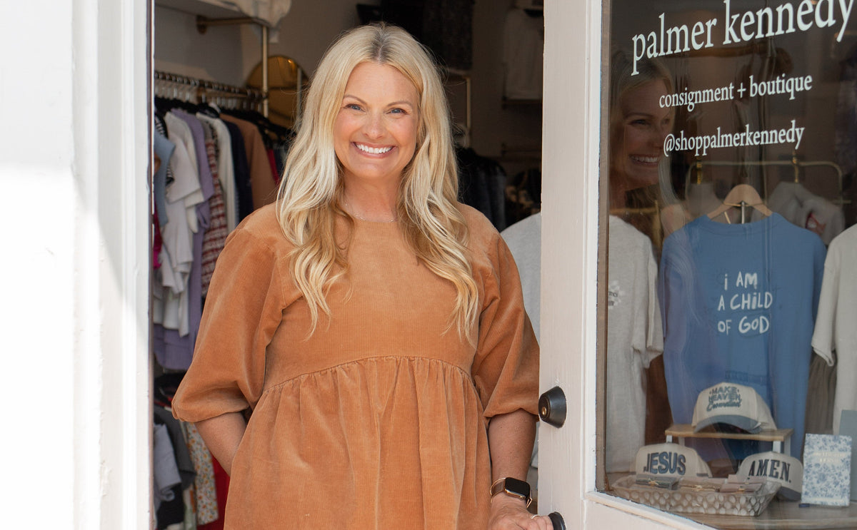 A woman stands in the doorway of a boutique wearing a tan corduroy dress and white cowboy boots. The white door has a glass window displaying the brand name 'palmer kennedy boutique'.